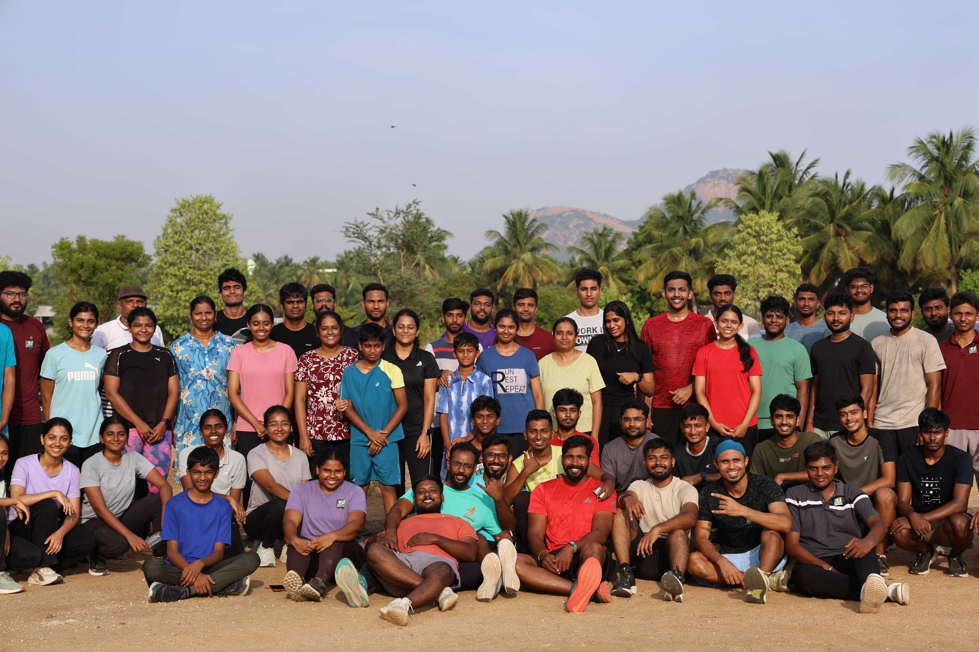 The full RUN 34 club gathered on open ground at sunrise - runners of every age standing and seated in rows, with palm trees and the Tiruchengode hills behind them.